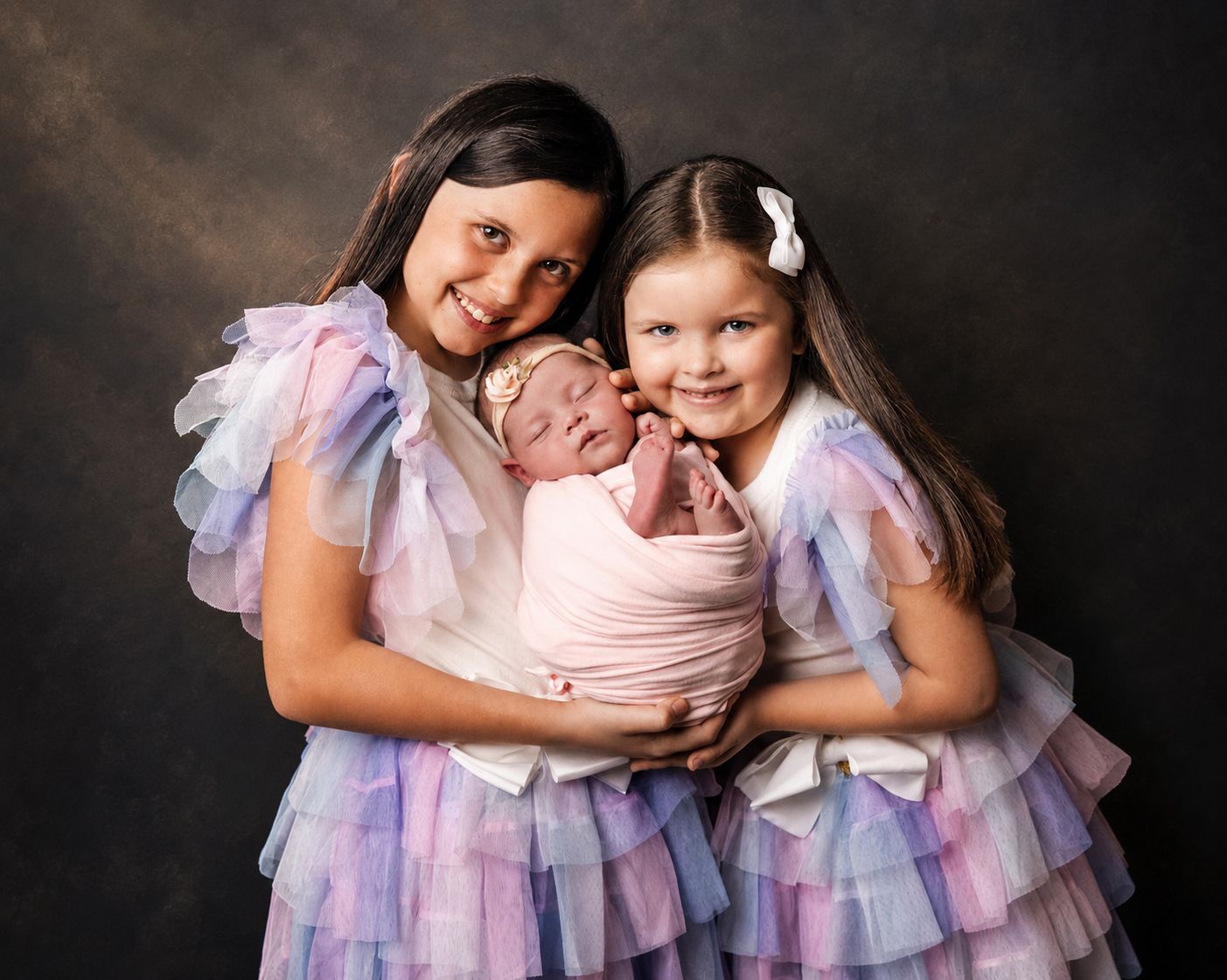 Two sisters in matching rainbow tulle dresses lovingly hold their swaddled newborn sibling for a professional portrait.