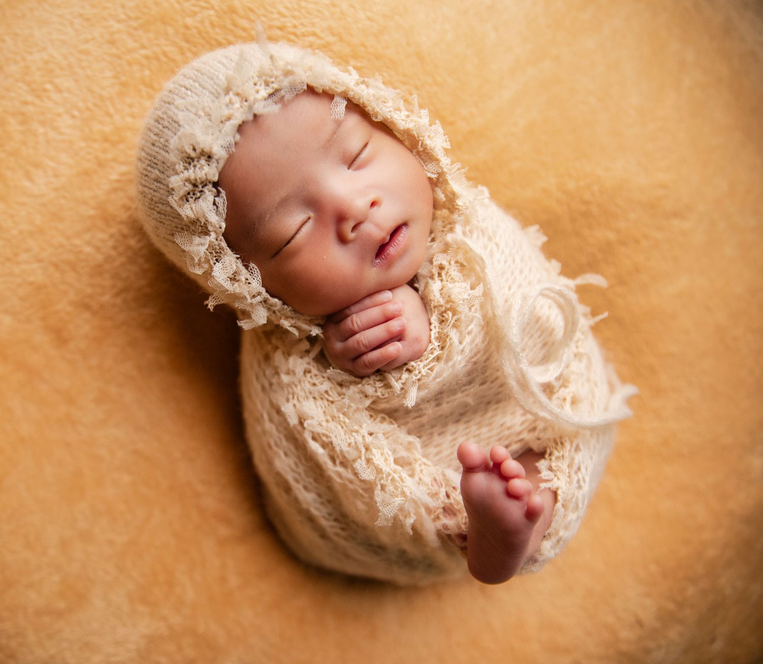 Sleeping newborn baby wrapped in cream lace swaddle wearing a matching bonnet on a soft tan background.