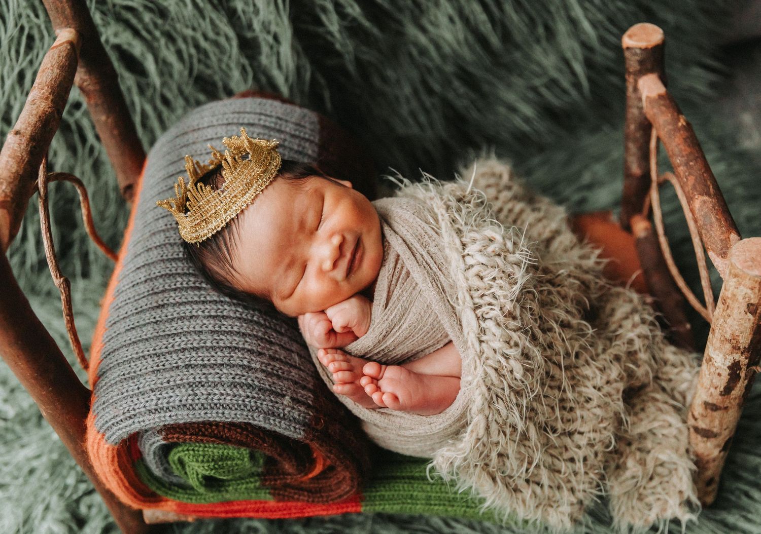 Smiling newborn baby wearing a tiny gold crown, swaddled in a gray wrap, resting on a rustic wooden bed with fur blanket.