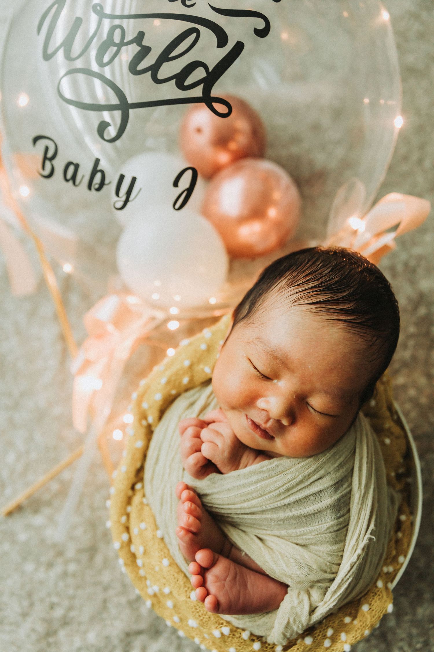 Newborn baby sleeping in a golden bucket wrapped in white, beside a balloon reading Baby J.