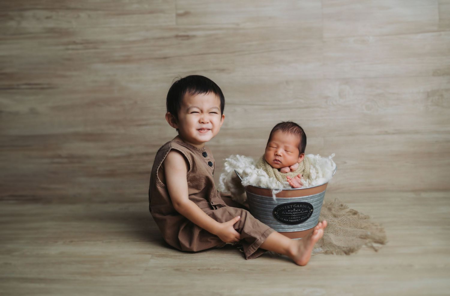 Smiling toddler boy sits beside a newborn baby sleeping in a small bucket during a sibling newborn photo session.