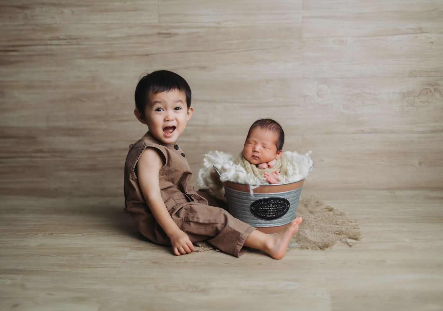Toddler boy in brown overalls sits beside a newborn baby in a decorative bucket on a wooden floor.