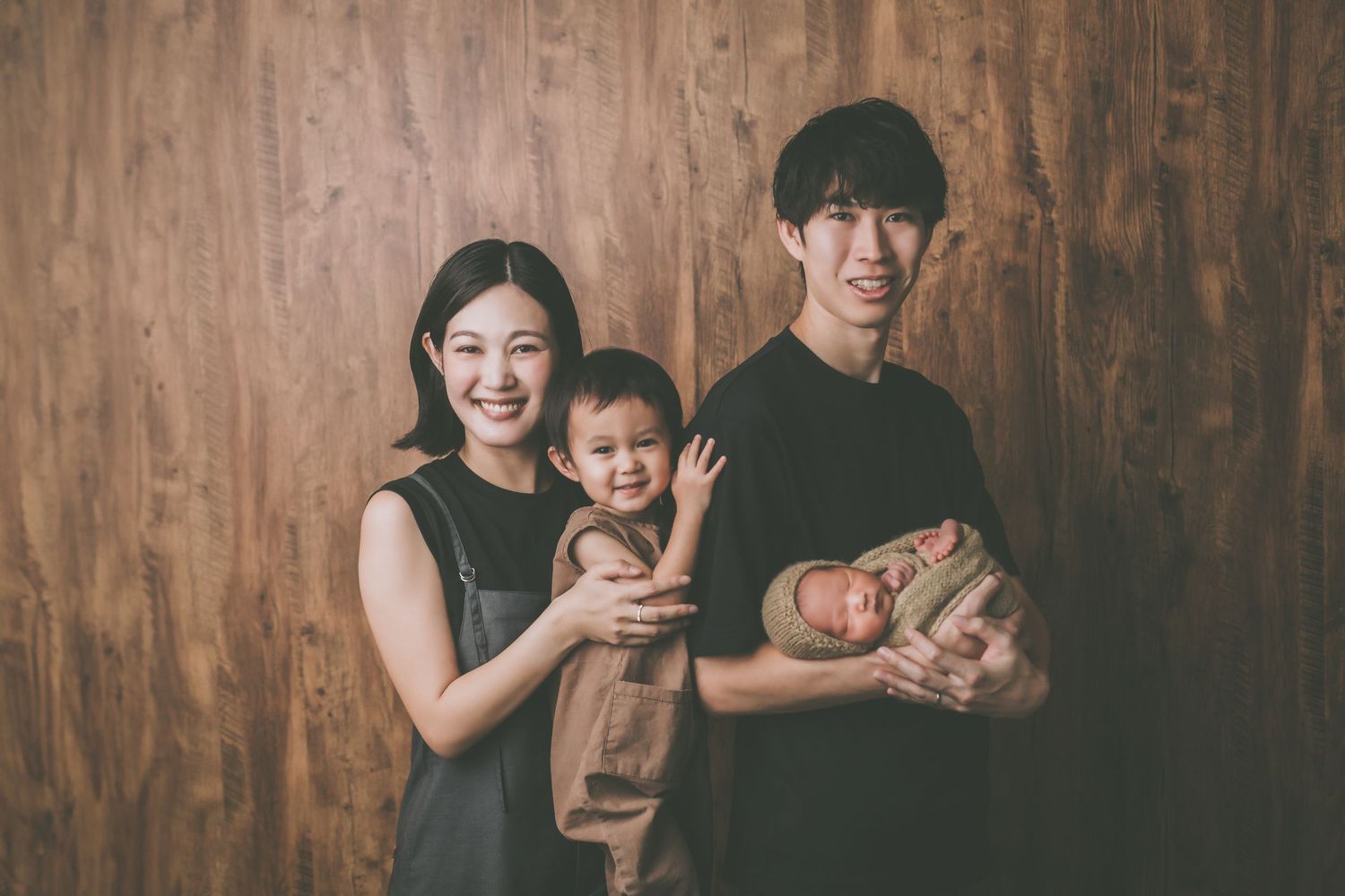 A young Asian family poses warmly, parents holding a toddler and newborn baby against a wooden backdrop.