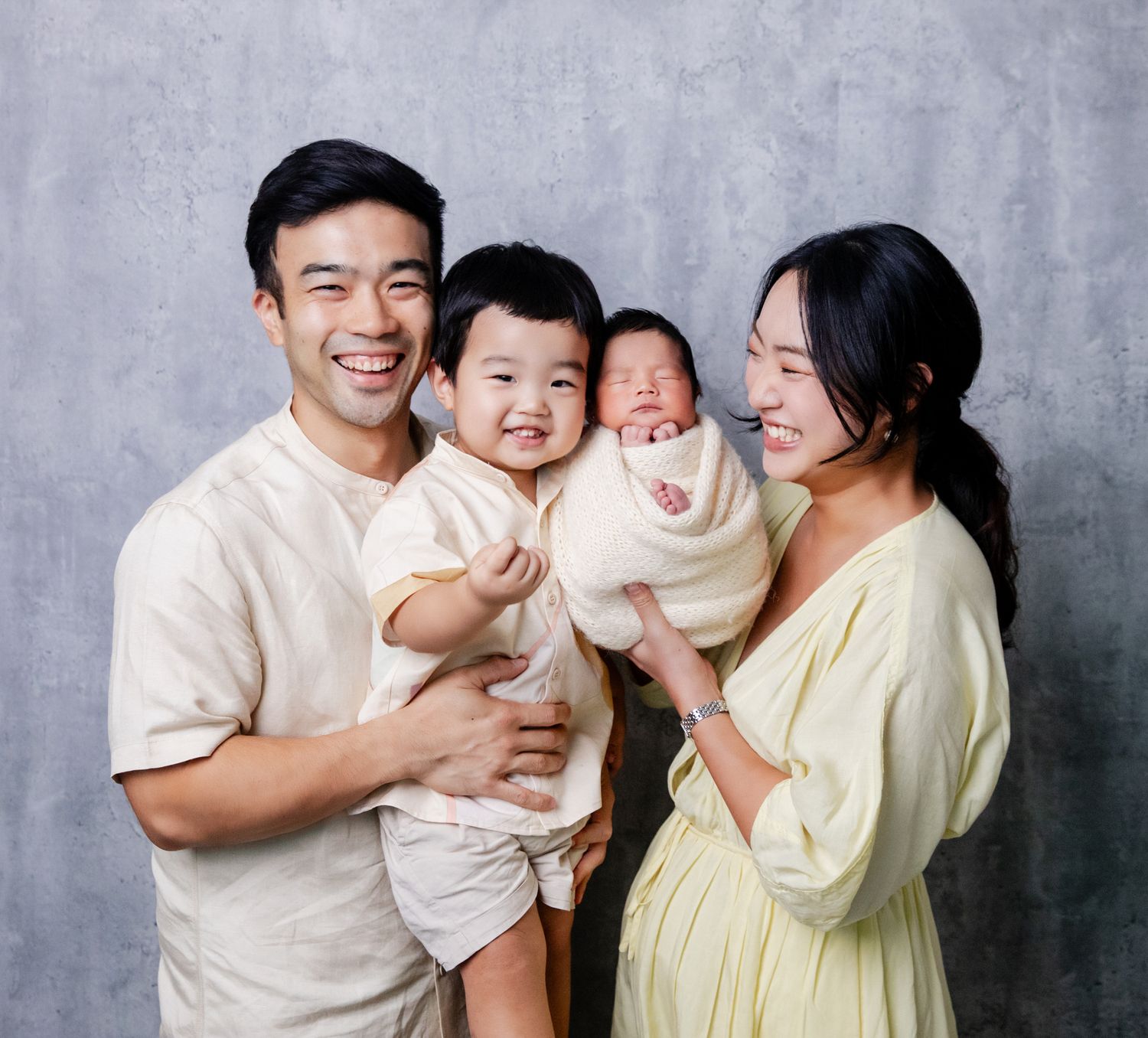 Happy Asian family portrait with parents holding toddler and newborn baby against gray background.