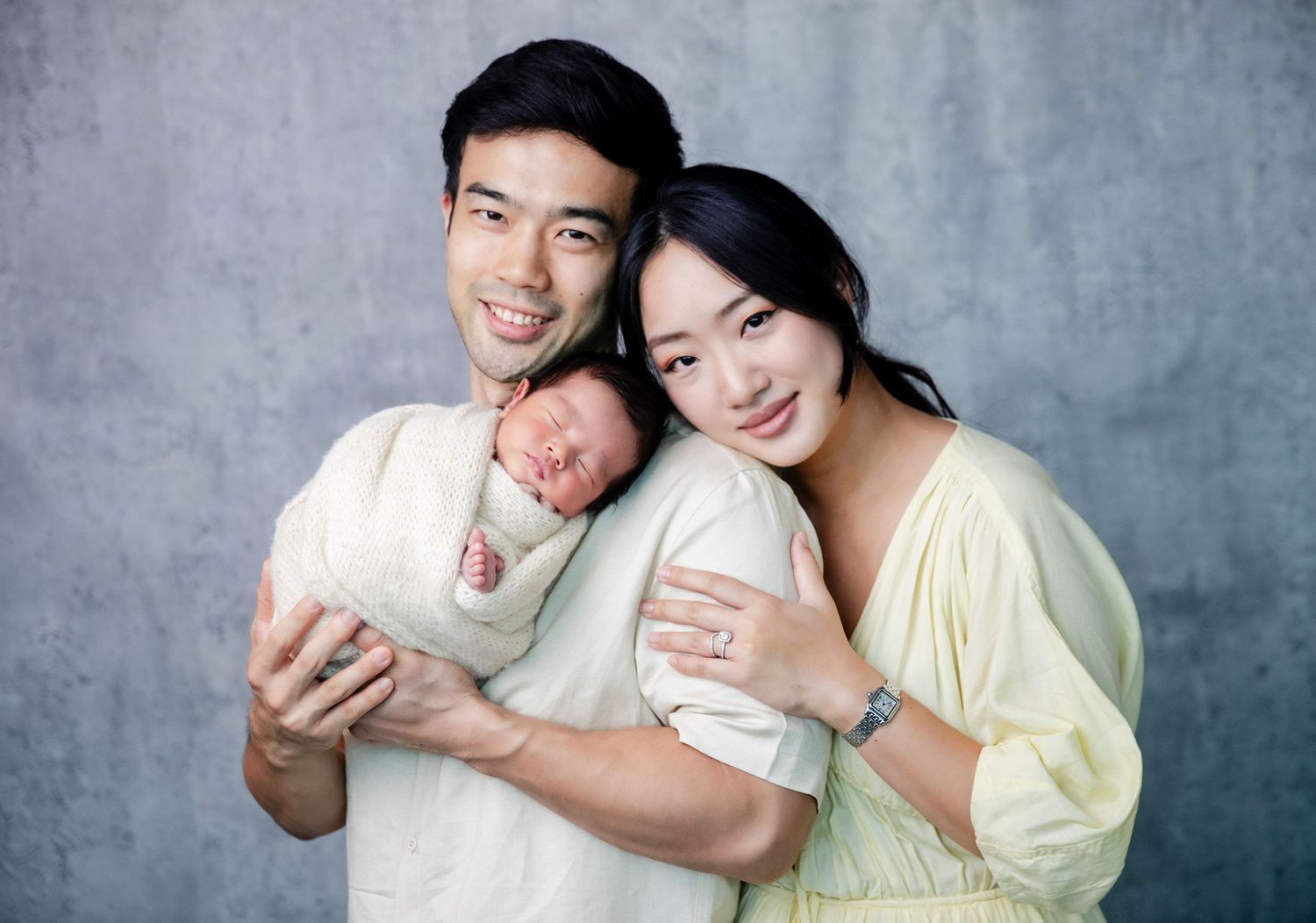 Asian couple smiling and holding their newborn baby wrapped in white, posing for a professional family portrait.