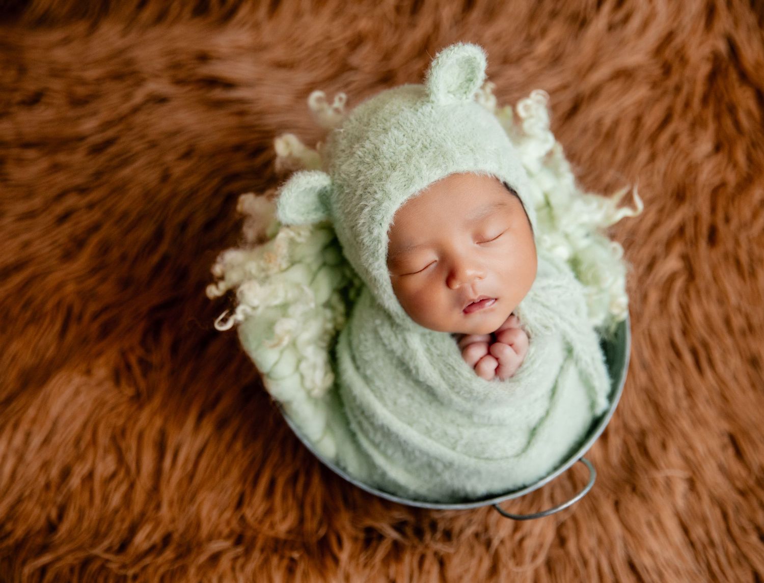 Newborn baby swaddled in mint green wrap and bear bonnet sleeping peacefully in a small bucket on brown fur.