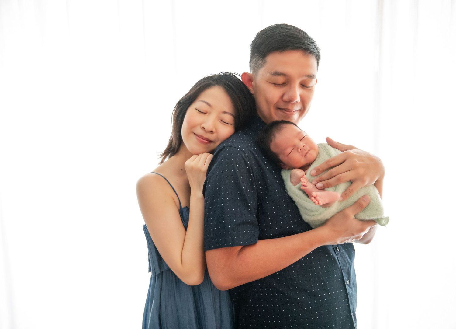 Happy Asian parents smiling and cuddling their sleeping newborn baby wrapped in a soft blanket.