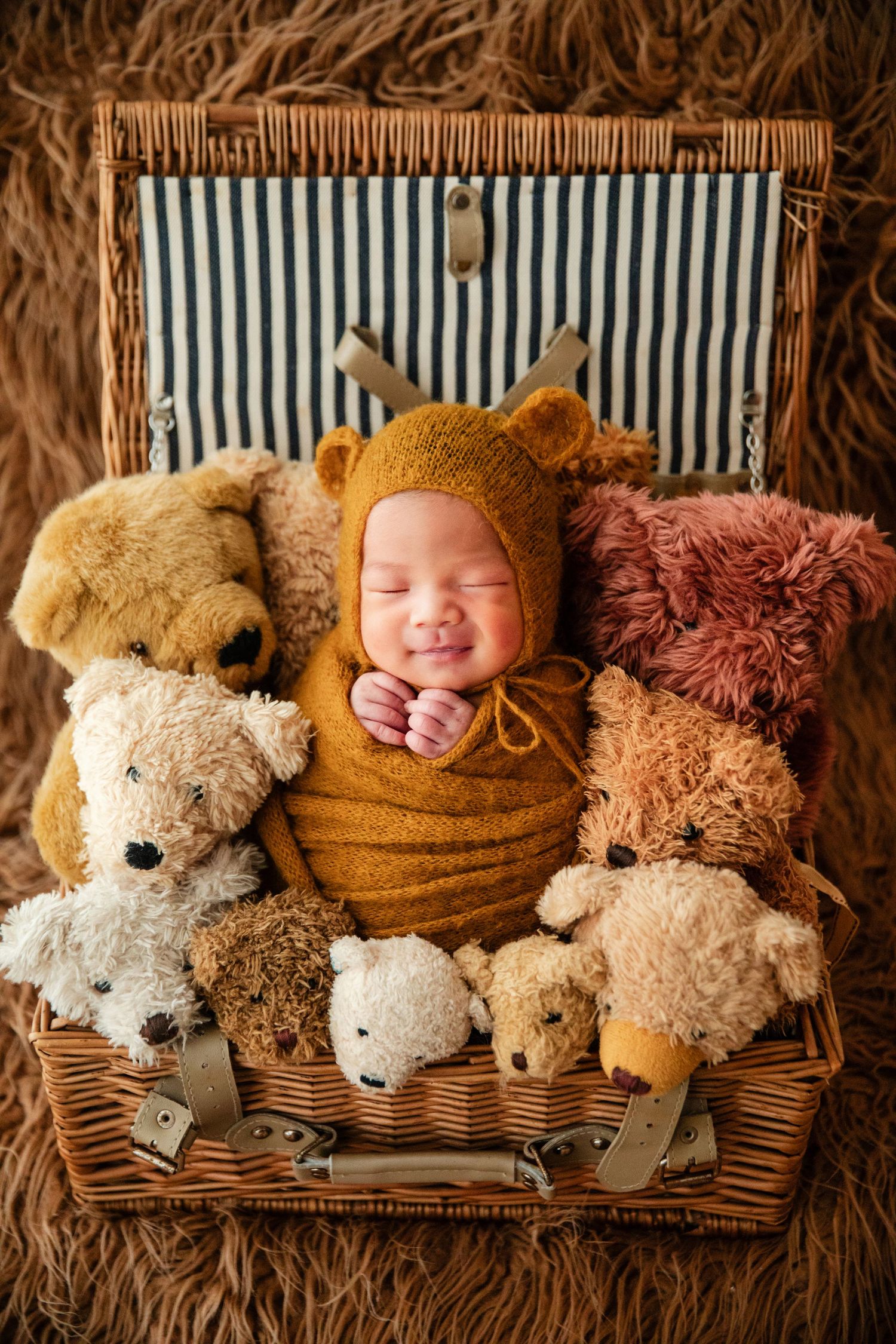 Smiling newborn in bear bonnet wrapped in mustard swaddle, nestled among teddy bears in a wicker picnic basket.