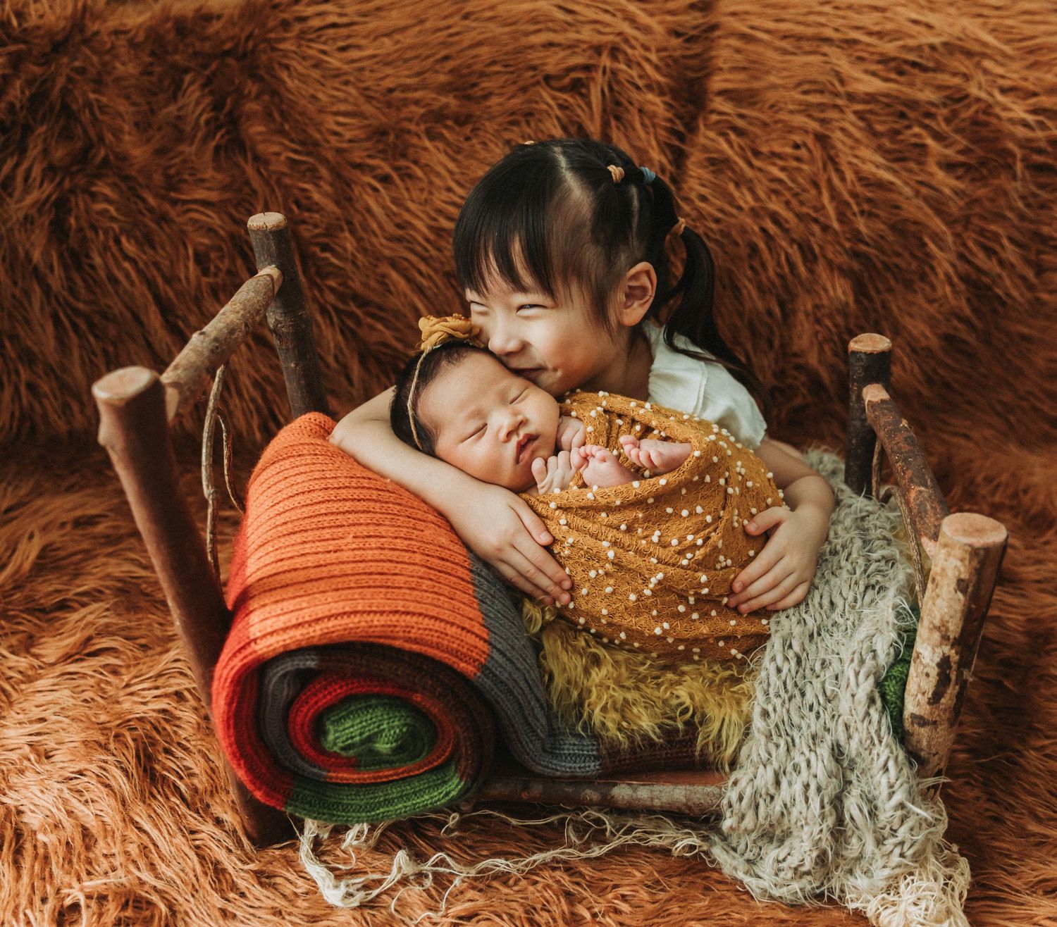 A young girl lovingly kisses a sleeping newborn wrapped in yellow, nestled together on a rustic wooden chair with warm fur backdrop.