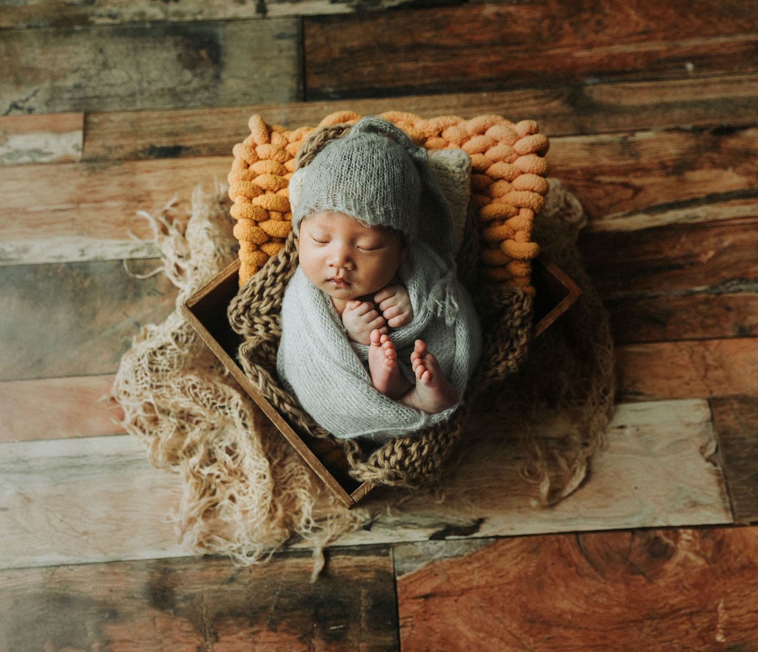 Newborn baby sleeping in a wooden box with a gray knit bonnet, wrapped in a soft blanket on a rustic wood floor.