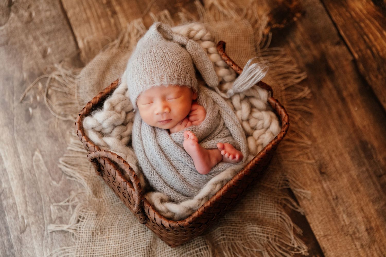 Sleeping newborn baby in gray knit outfit and hat nestled in a wicker basket with soft wool blanket.