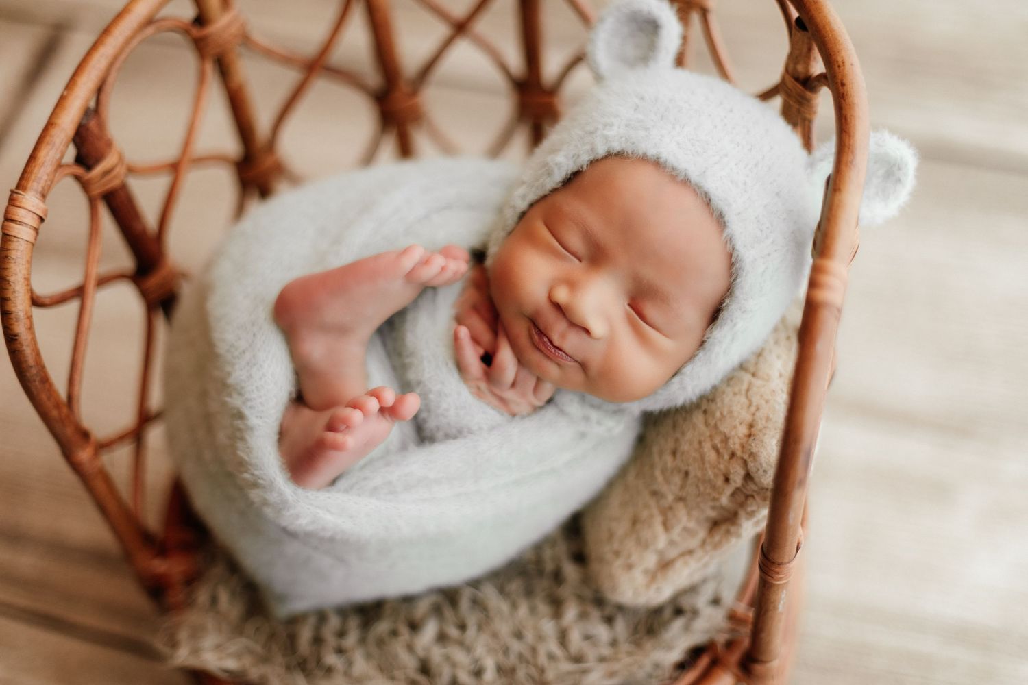 Smiling newborn baby wrapped in soft gray blanket wearing bear bonnet, sleeping peacefully in wicker basket.