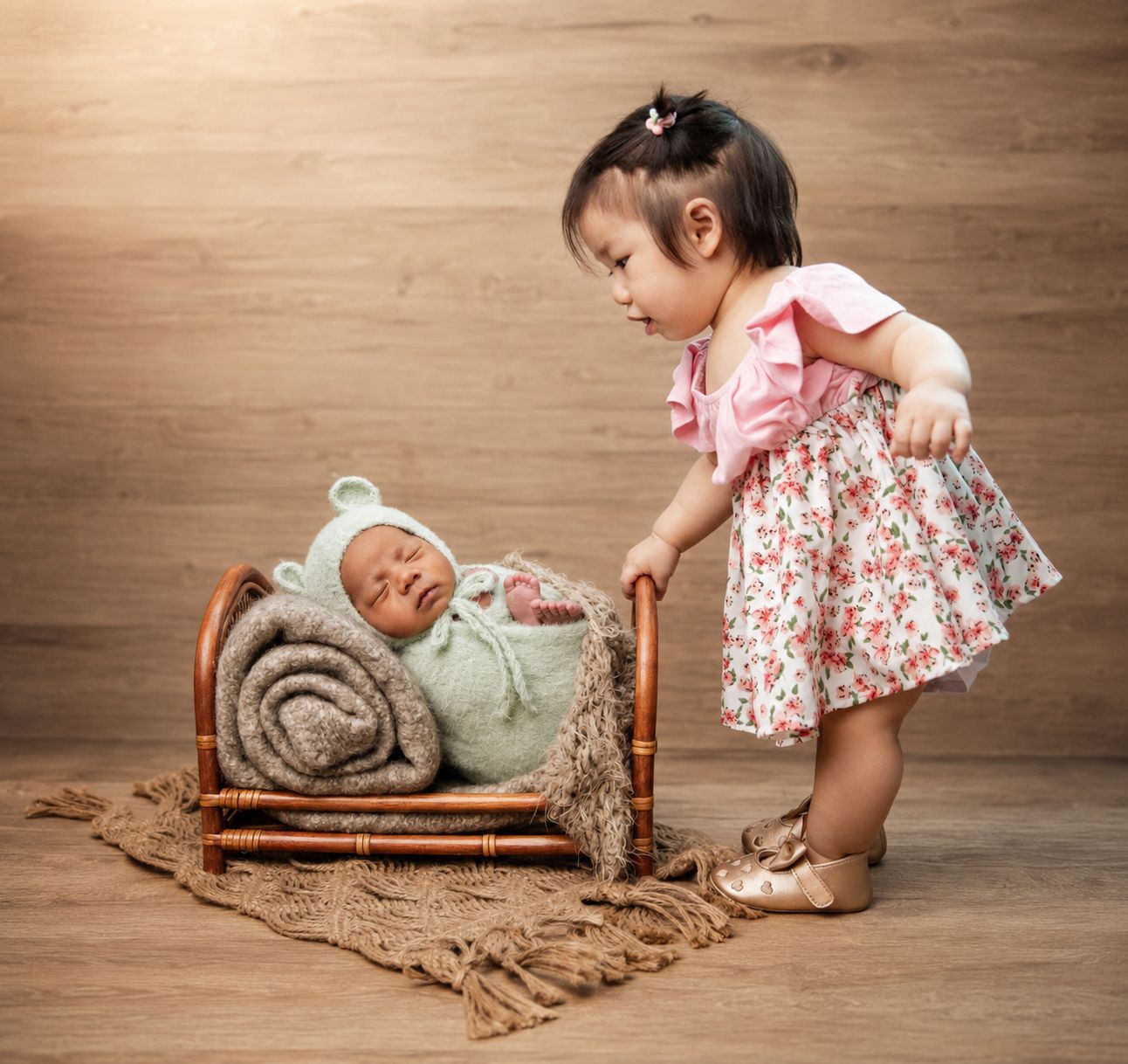 Toddler girl in floral dress admiring sleeping newborn baby wrapped in green knit in a small wooden bed.