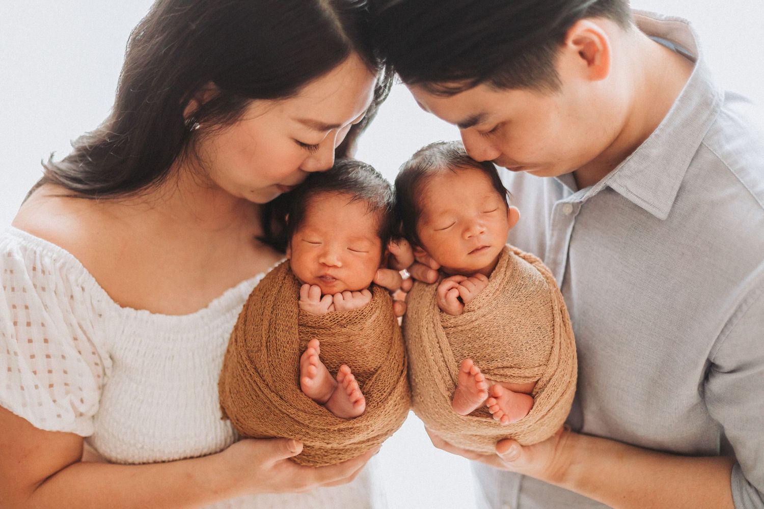 Asian couple holding twin newborns wrapped in brown swaddles during a professional newborn photography session.