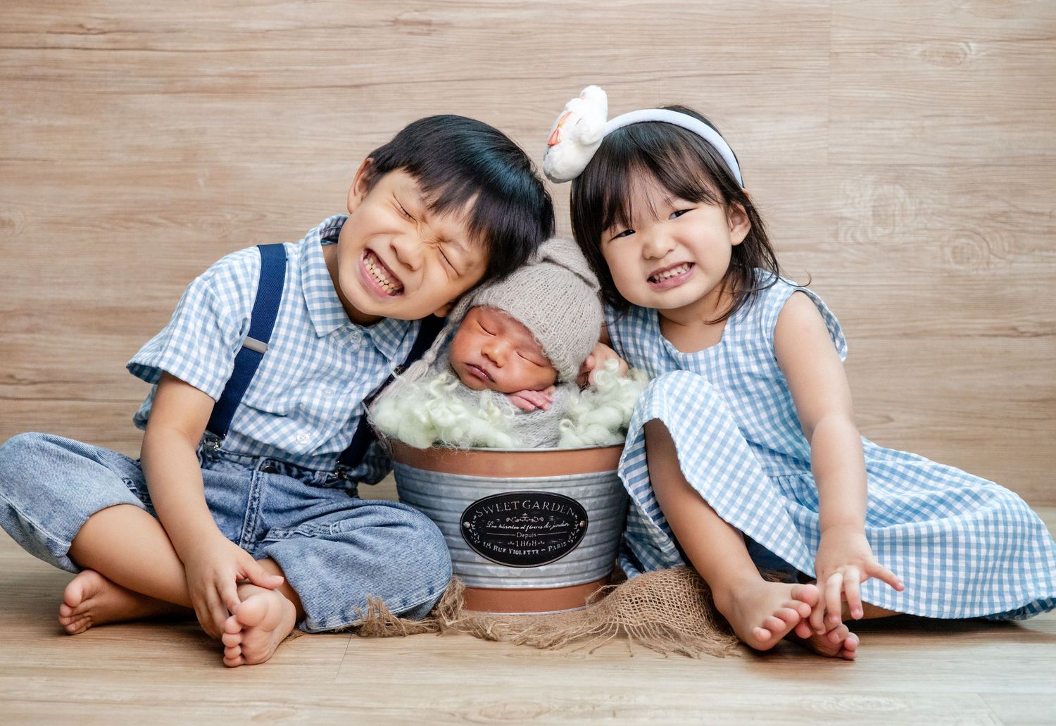 Two smiling Asian children pose with a sleeping newborn baby in a decorative bucket for a sibling portrait photo.