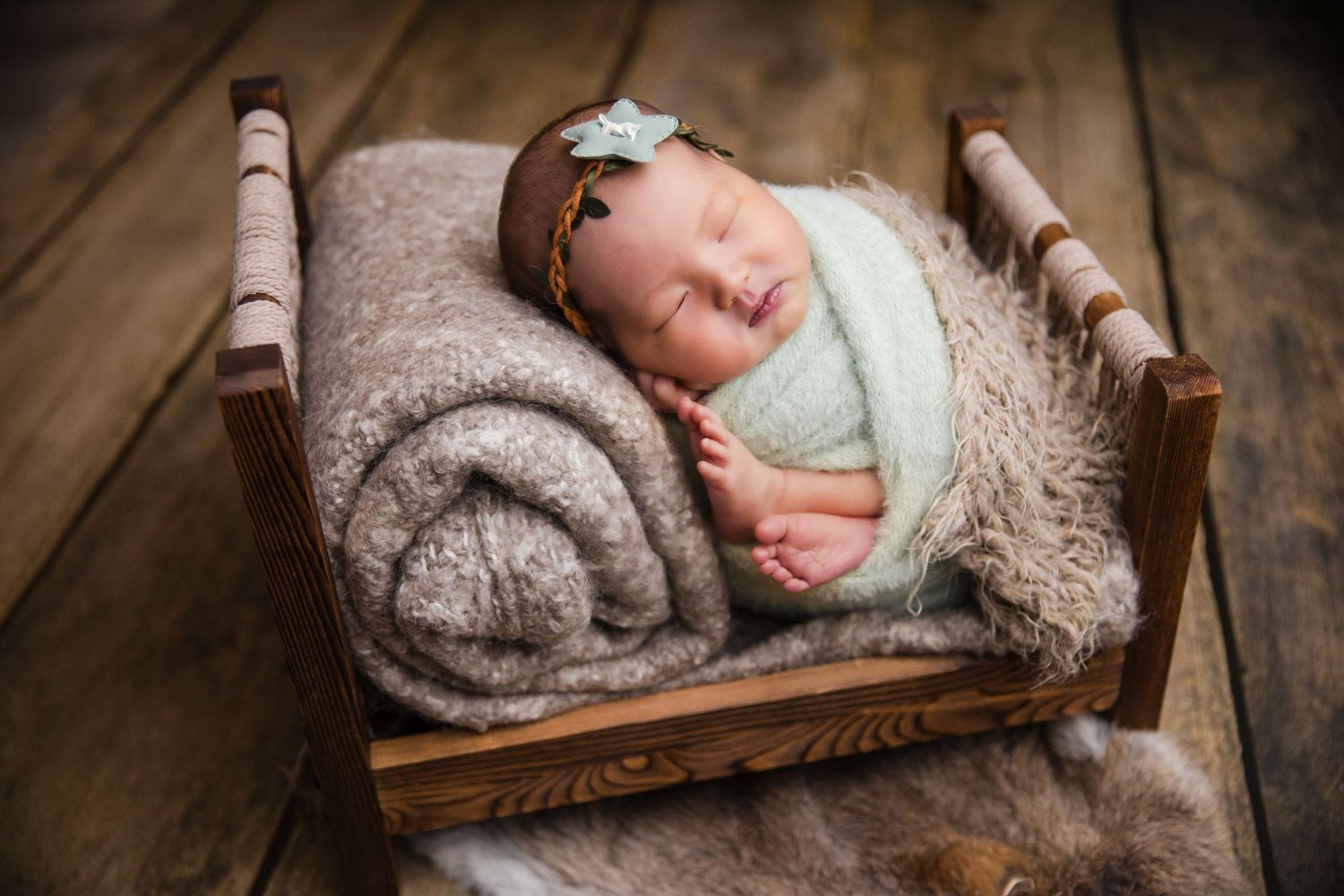 Newborn baby sleeping peacefully in a rustic wooden mini bed with soft blankets and a floral headband.