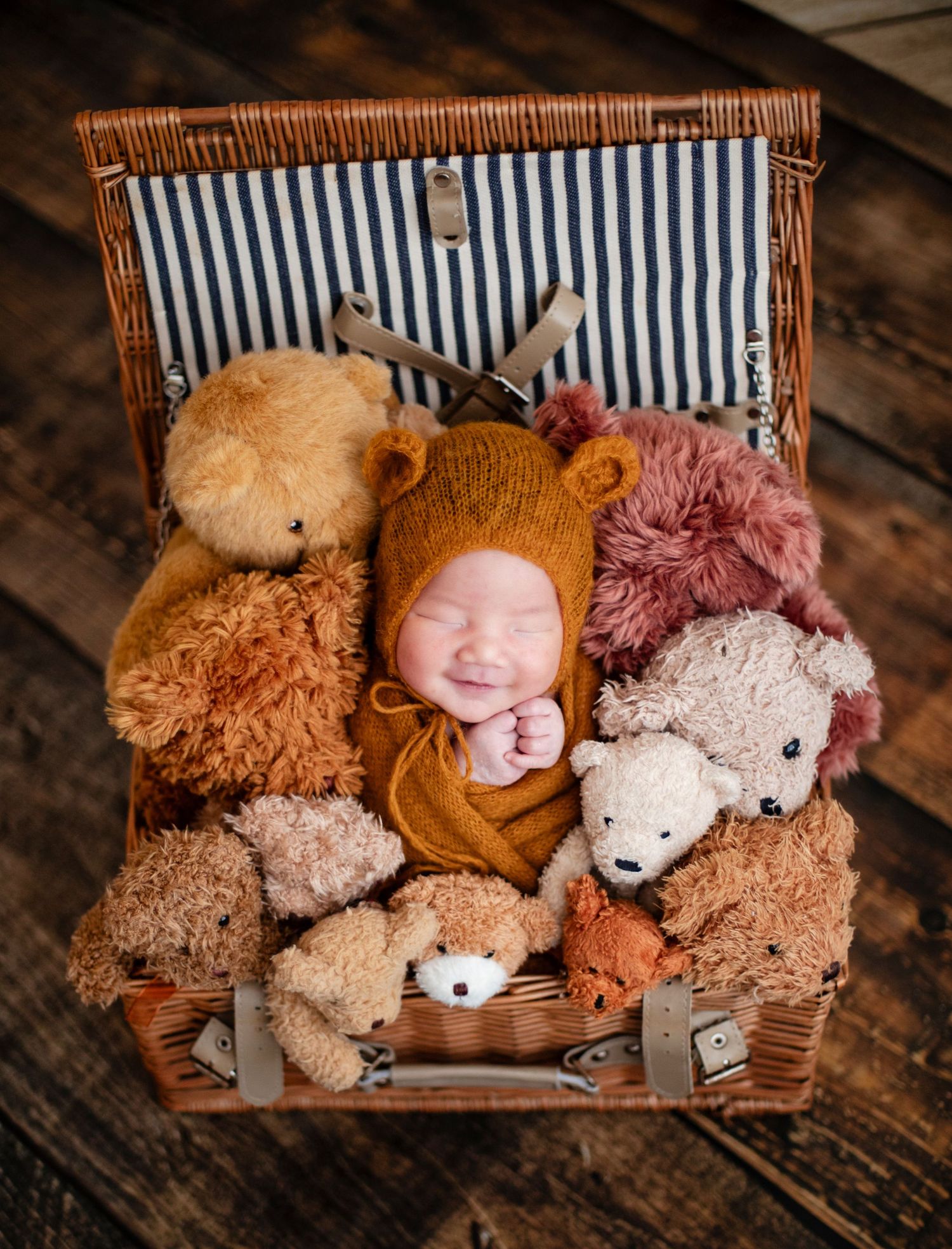 Smiling newborn in mustard bear bonnet surrounded by teddy bears inside an open wicker suitcase.