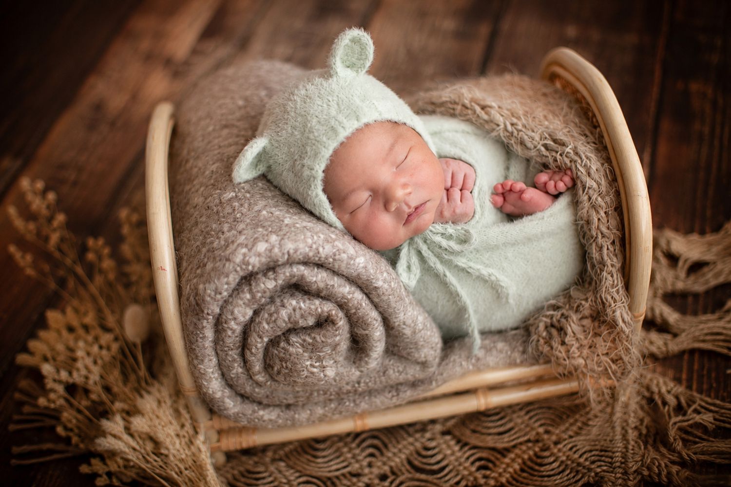 Newborn baby sleeping peacefully in a tiny wooden bed, wrapped in a mint green knit blanket and bear ear bonnet.