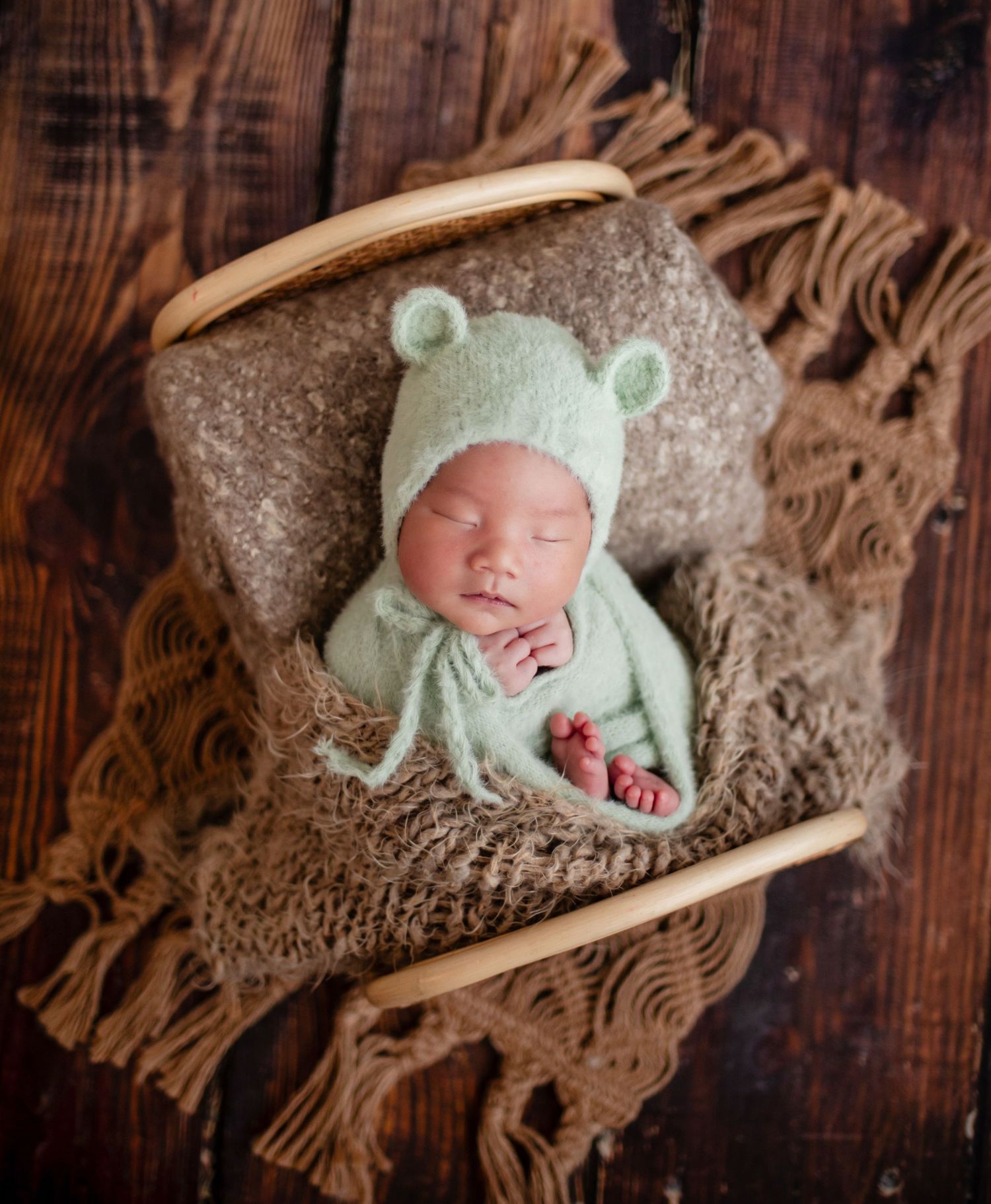 Sleeping newborn in green bear bonnet and outfit nestled in wooden bowl with brown knit blanket.