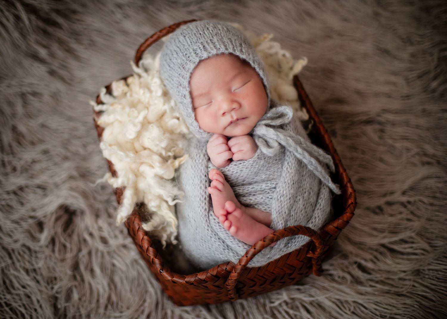 Newborn baby sleeping peacefully in a wicker basket, wrapped in a gray knit swaddle and bonnet on a soft fur blanket.