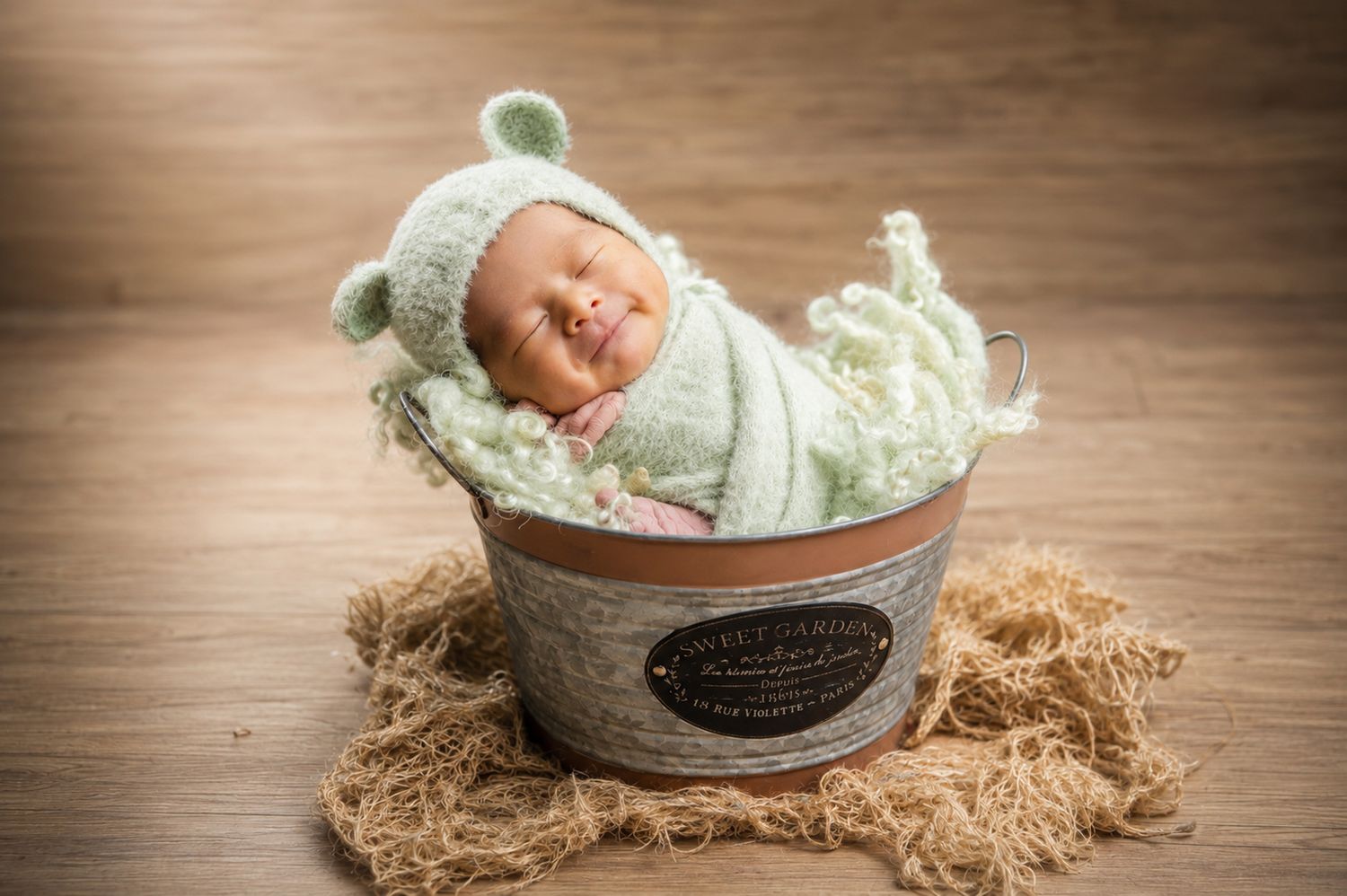 Newborn baby sleeping in a rustic metal bucket, wrapped in mint green, wearing a cozy bear hat.