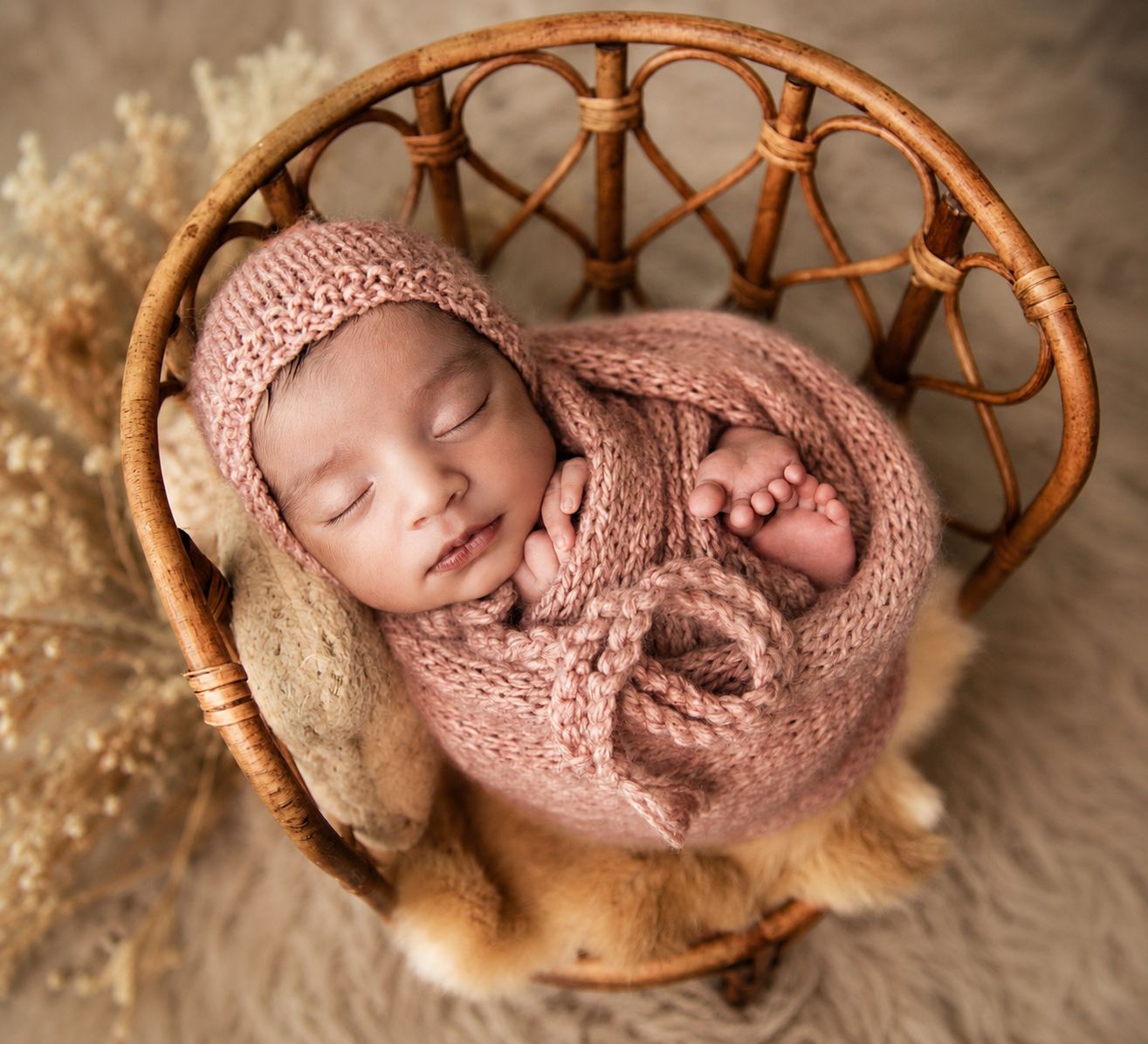 Newborn baby sleeping peacefully in a wicker basket, wrapped in a pink knit blanket and bonnet.
