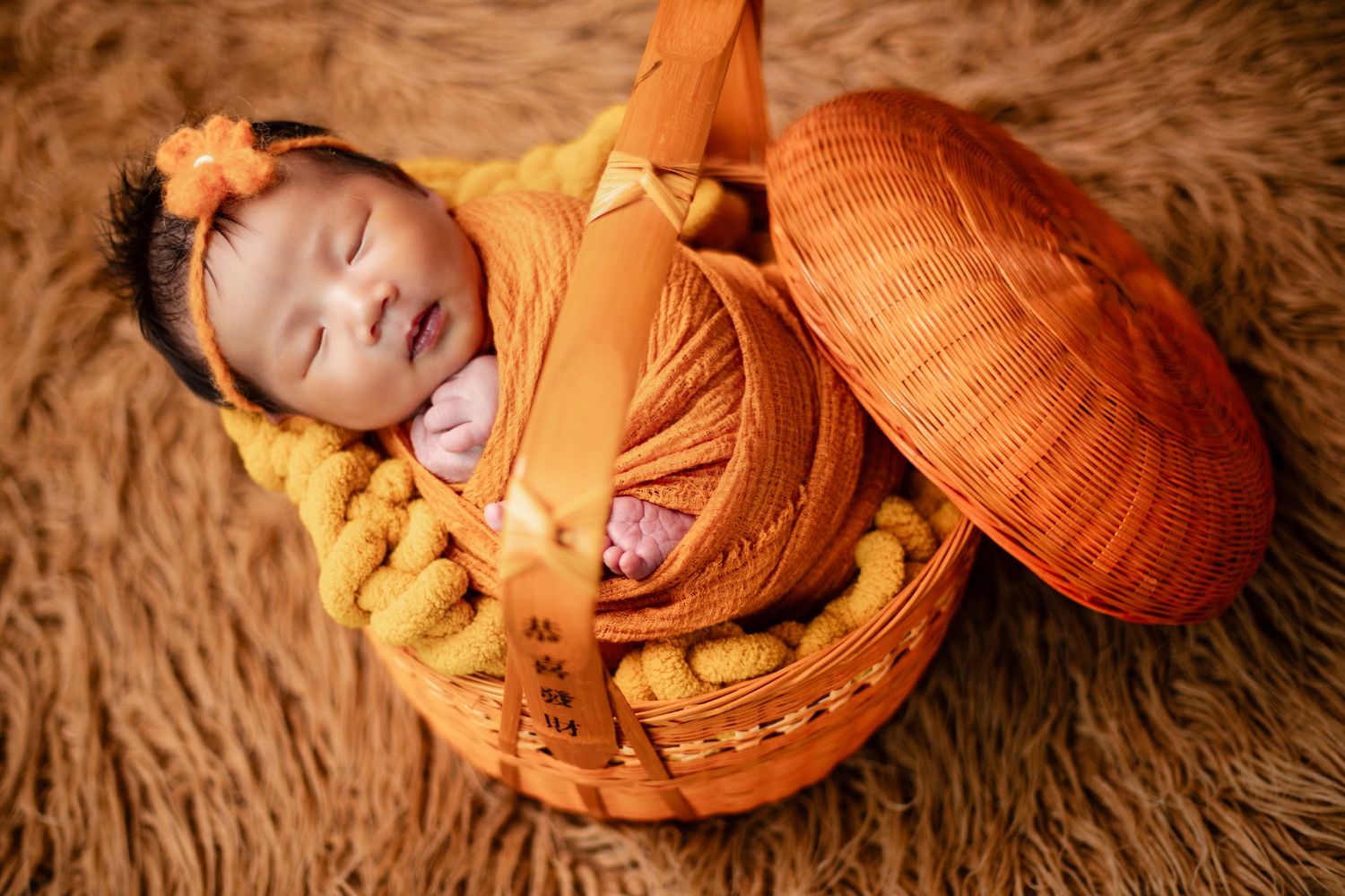 Newborn baby sleeping peacefully in an orange wicker basket, wrapped in orange fabric with a floral headband.