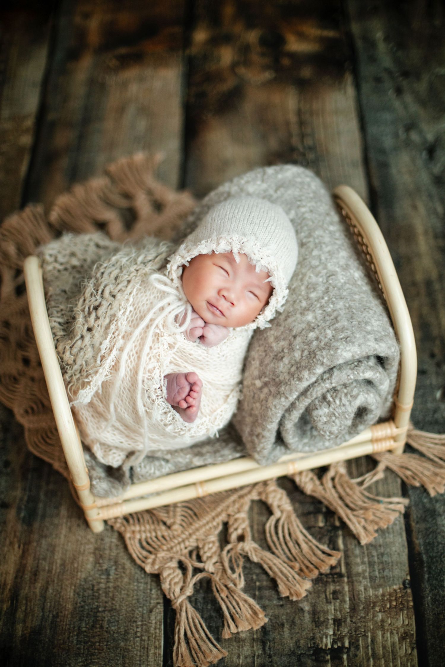 Sleeping newborn baby wrapped in white knit swaddle and bonnet resting in small wooden basket on rustic wood floor.
