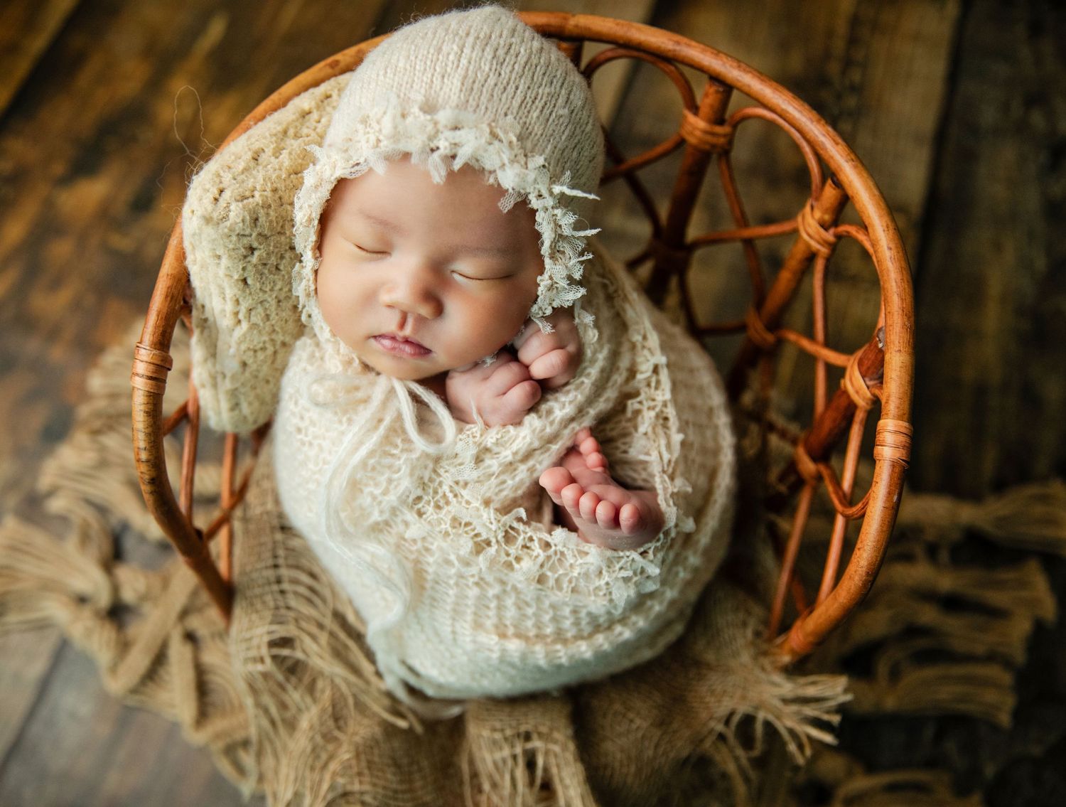 Sleeping newborn baby wrapped in cream knit swaddle and bonnet nestled in a wicker chair.