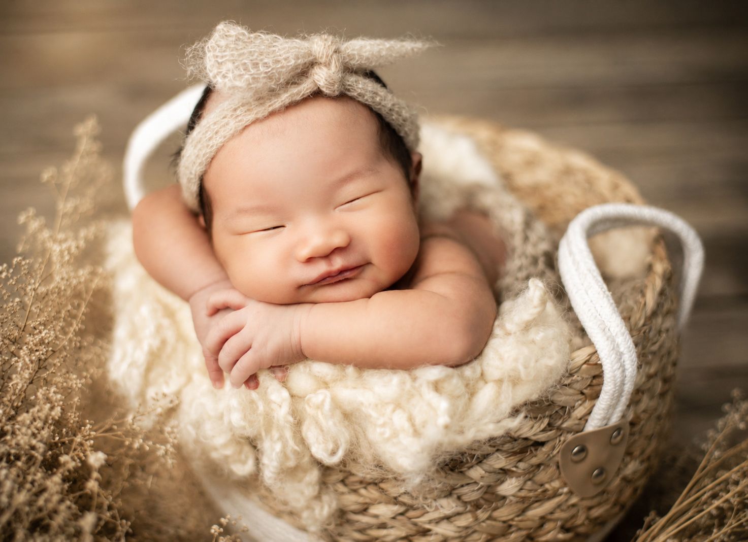 Smiling newborn baby in a knit bow hat, nestled in a woven basket with dried flowers.