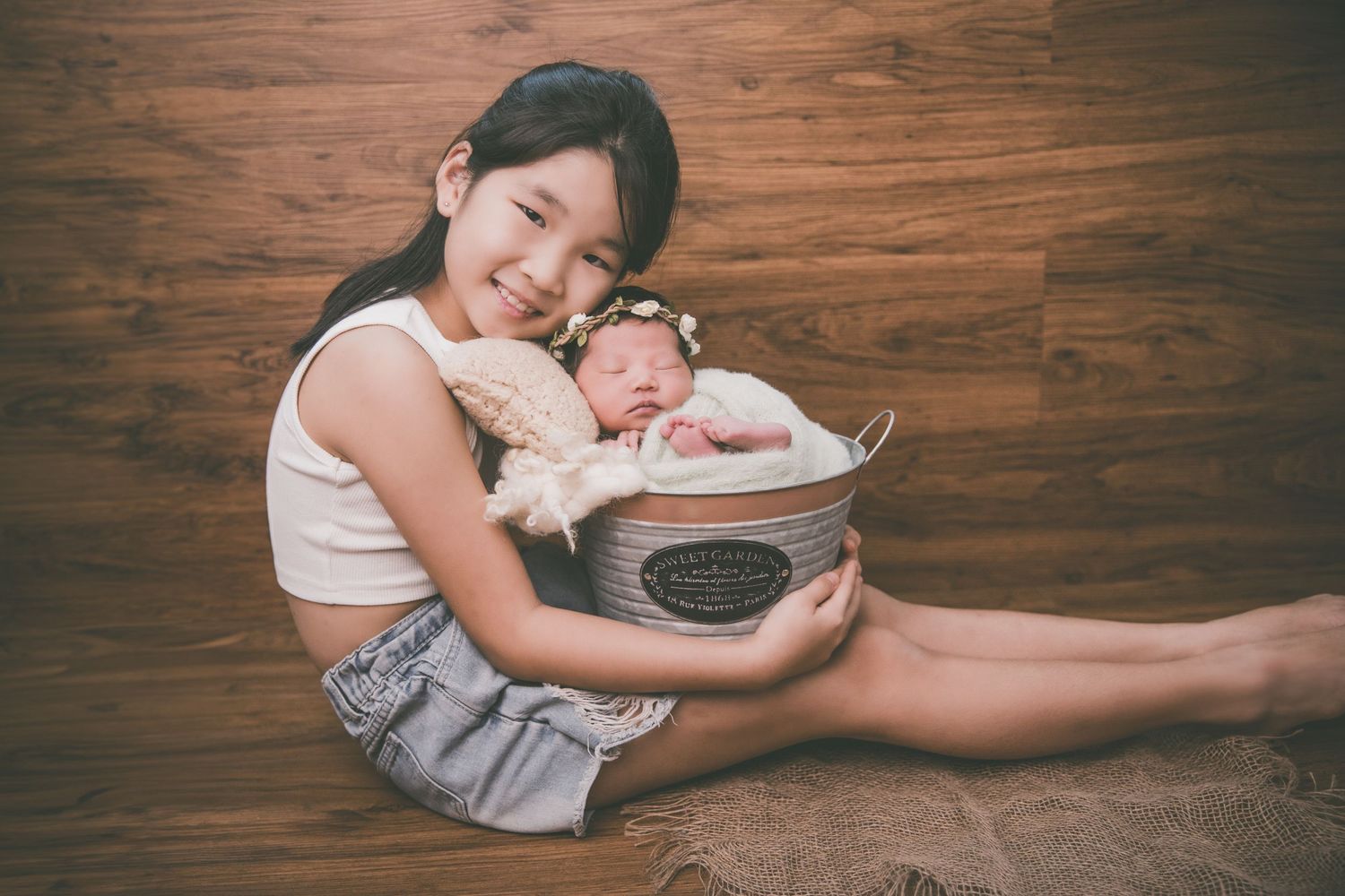 A young girl smiles while holding a wicker basket with a sleeping newborn baby on a wooden floor.
