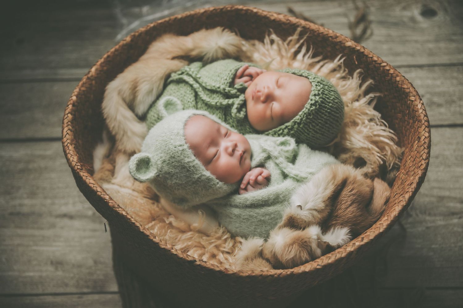 Two sleeping newborn twins in green knit bear outfits nestled in a rustic wicker basket with fur lining.