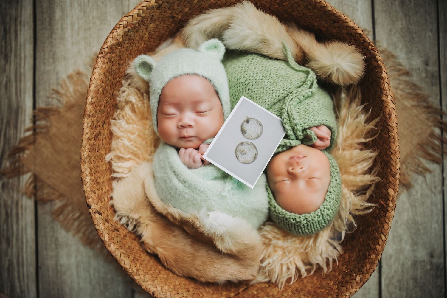 Two newborn twins sleeping in a wicker basket wearing green knit bear hats, wrapped in soft blankets with ultrasound photos.