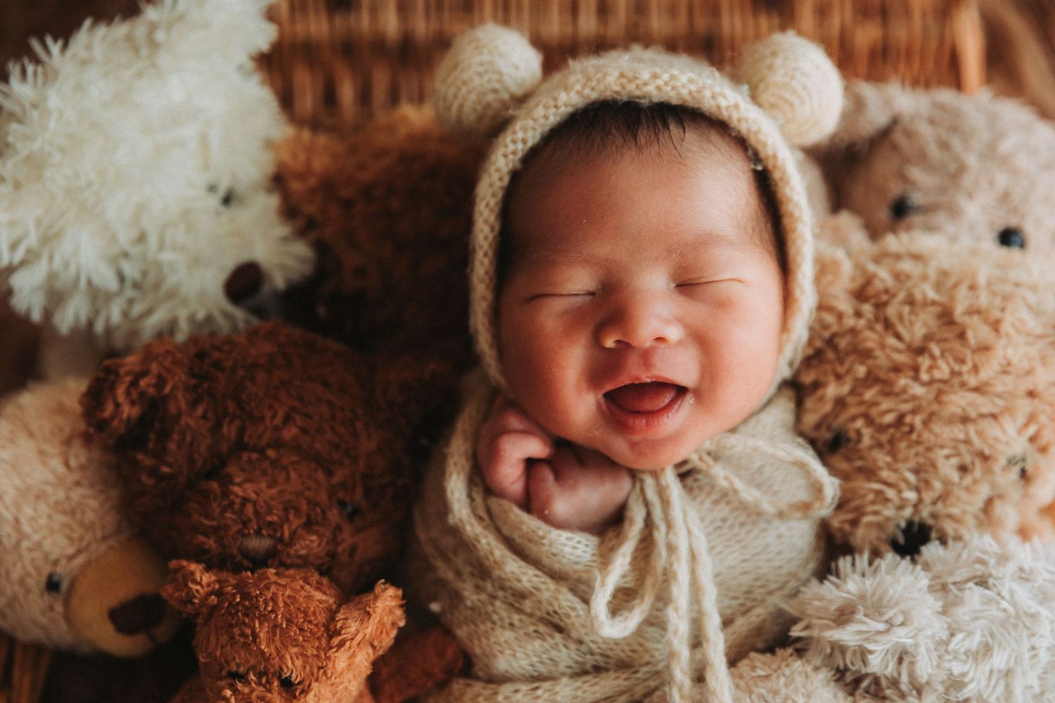 Smiling newborn baby in knit bear bonnet and wrap surrounded by teddy bears in a cozy basket.