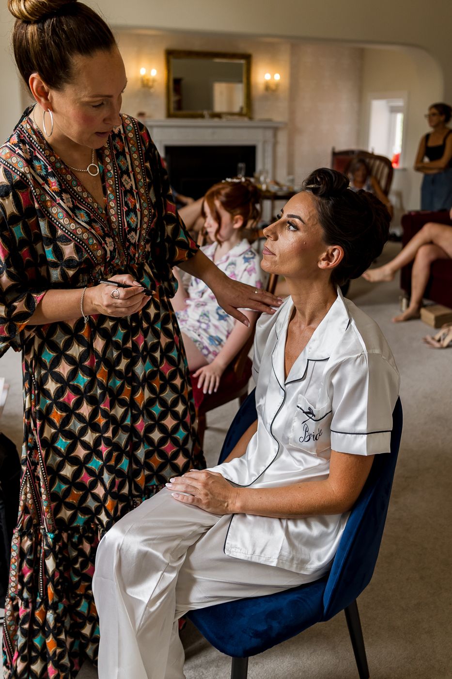 Makeup artist applying makeup to a bride in white satin pajamas seated on a blue chair in an elegant room.