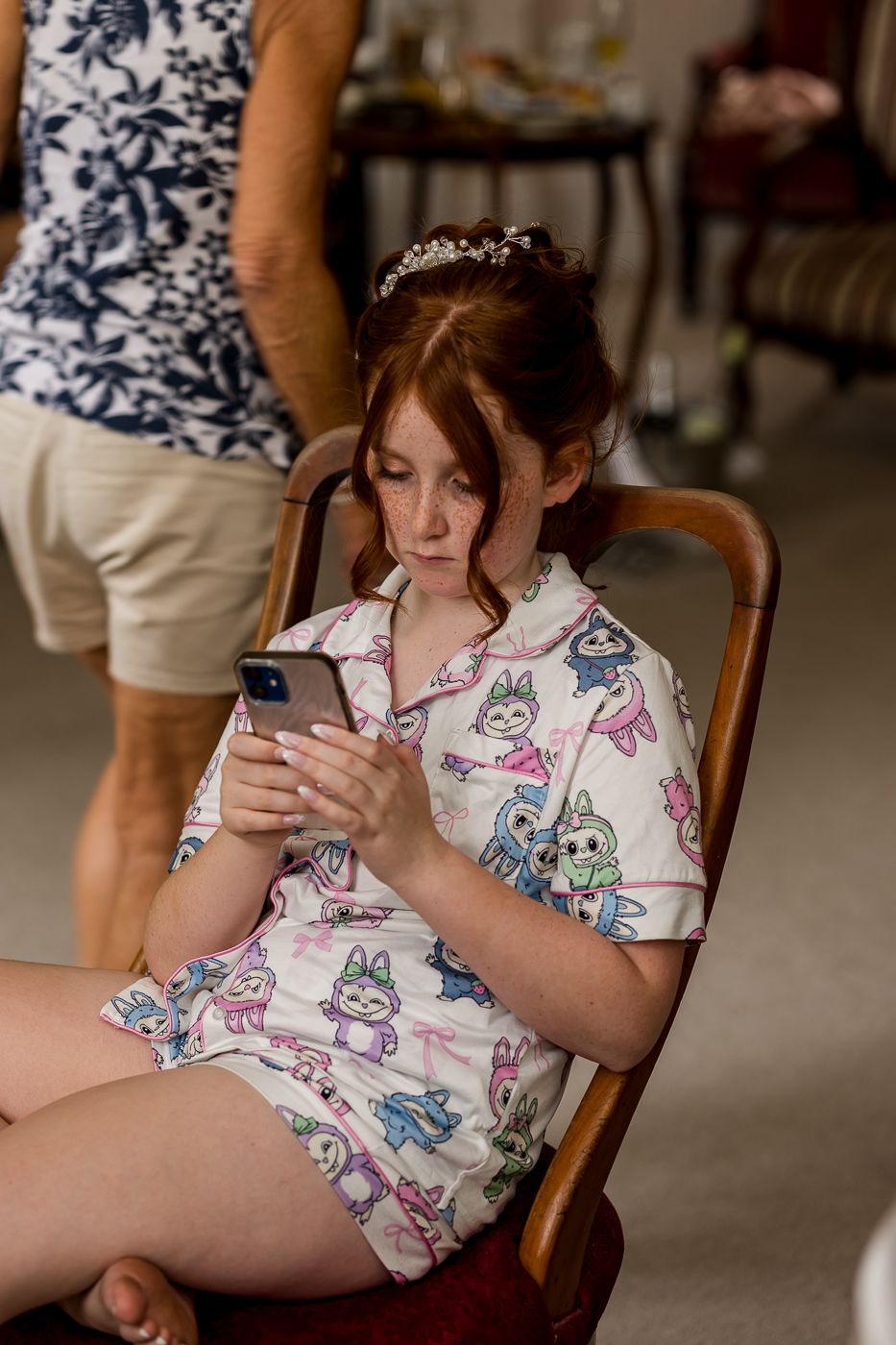 Young girl in unicorn pajamas wearing a tiara sits in wooden chair looking at smartphone.
