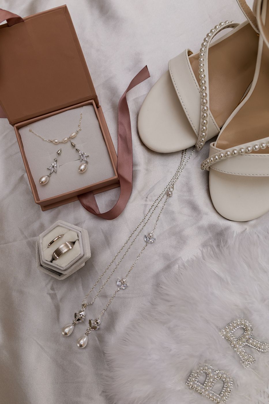 Elegant bridal accessories flatlay featuring pearl jewelry set, diamond necklaces, gold rings, and ivory heeled sandals on white fur.