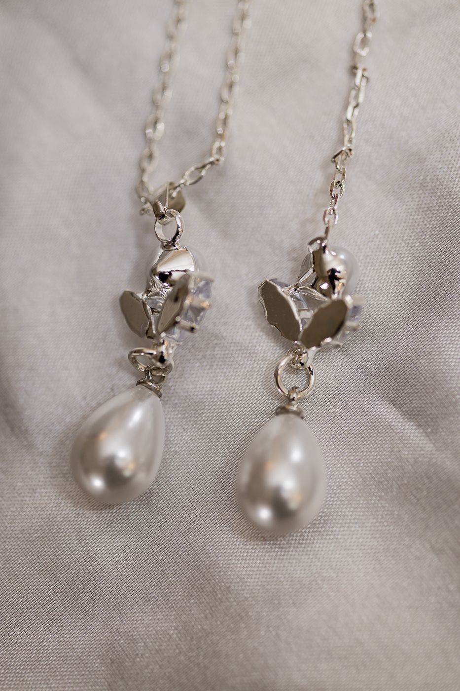 Silver chain earrings with crystal floral tops and teardrop pearl pendants displayed on soft gray fabric.