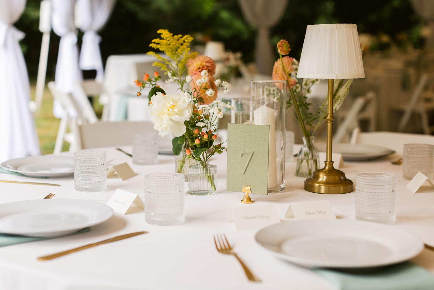 Elegant outdoor wedding reception table with peach and yellow flowers, brass lamp, table number card, plates and gold flatware.
