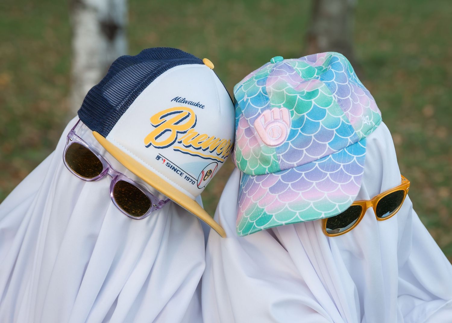 Two people in white ghost sheets wearing sunglasses, a Brewers baseball cap and a pastel mermaid scale cap, leaning together outdoors.
