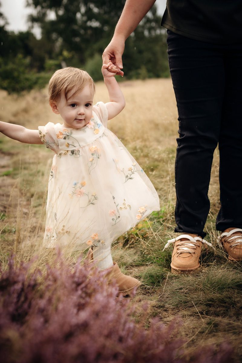 Toddler in a cream floral dress holds an adult's hand while walking through a grassy field with purple heather in the foreground.