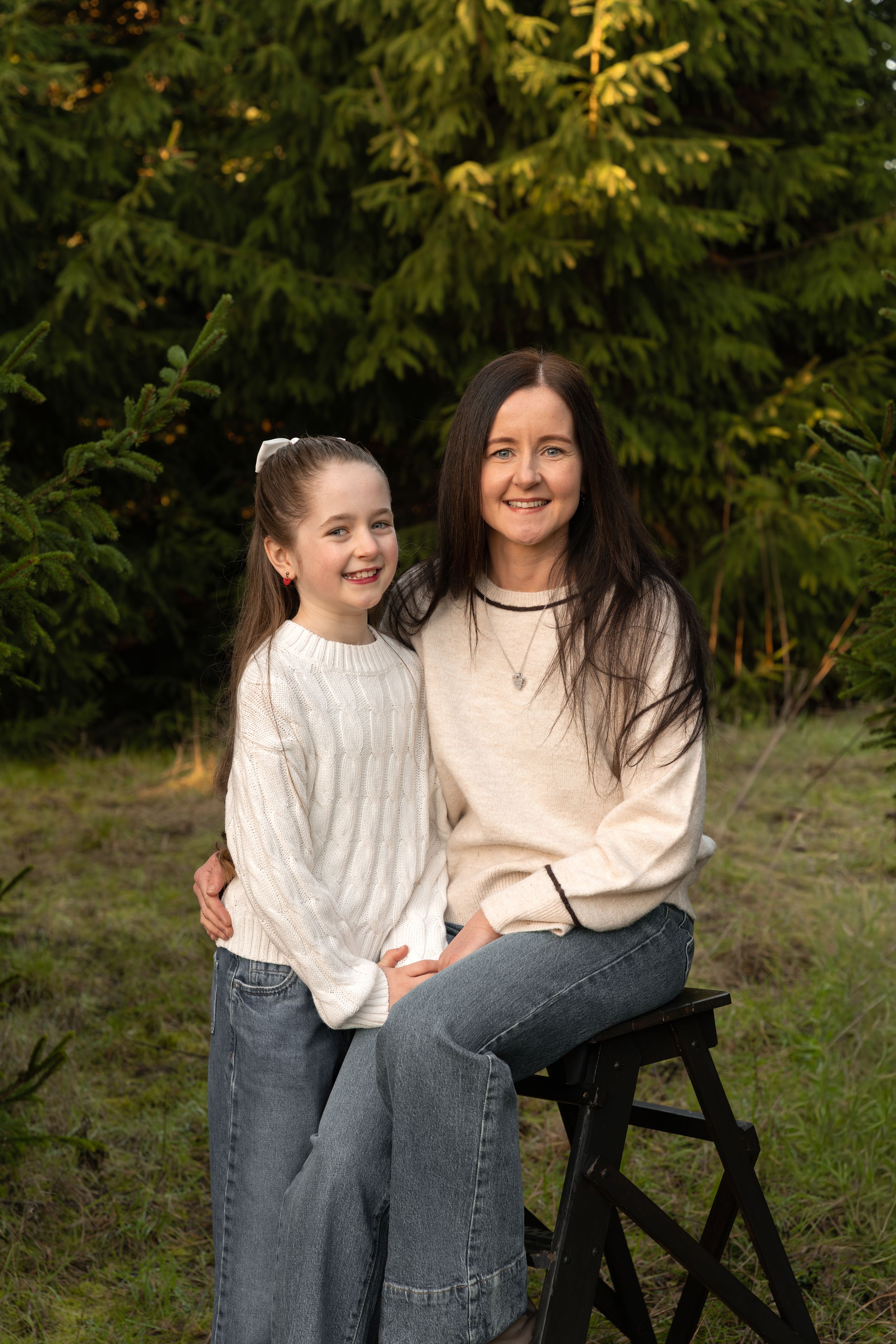 Smiling mother and daughter in cream sweaters and blue jeans pose together outdoors in front of green evergreen trees.