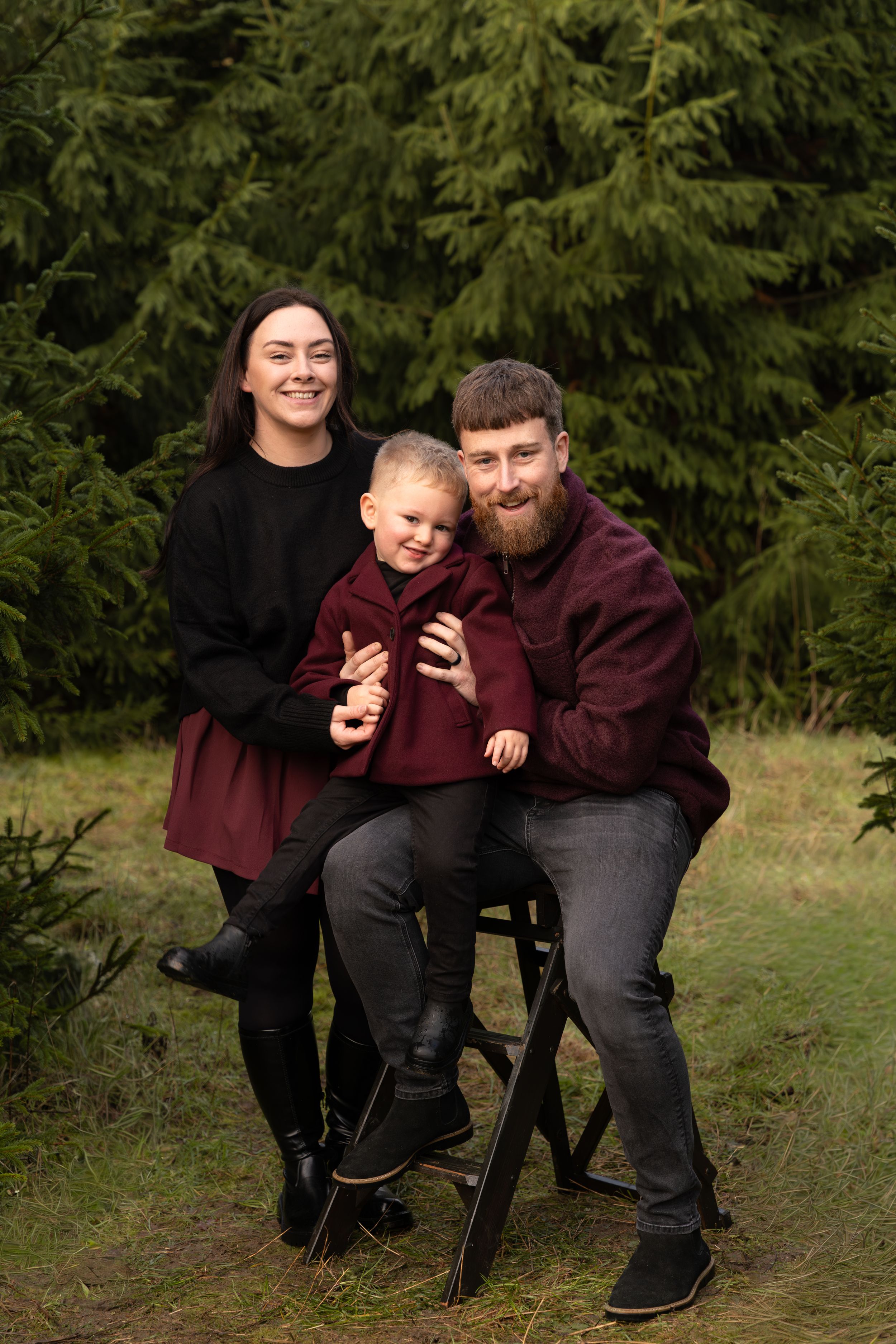 Smiling family of three in burgundy and black outfits poses outdoors, with a bearded man seated on a stool holding a toddler beside a woman.