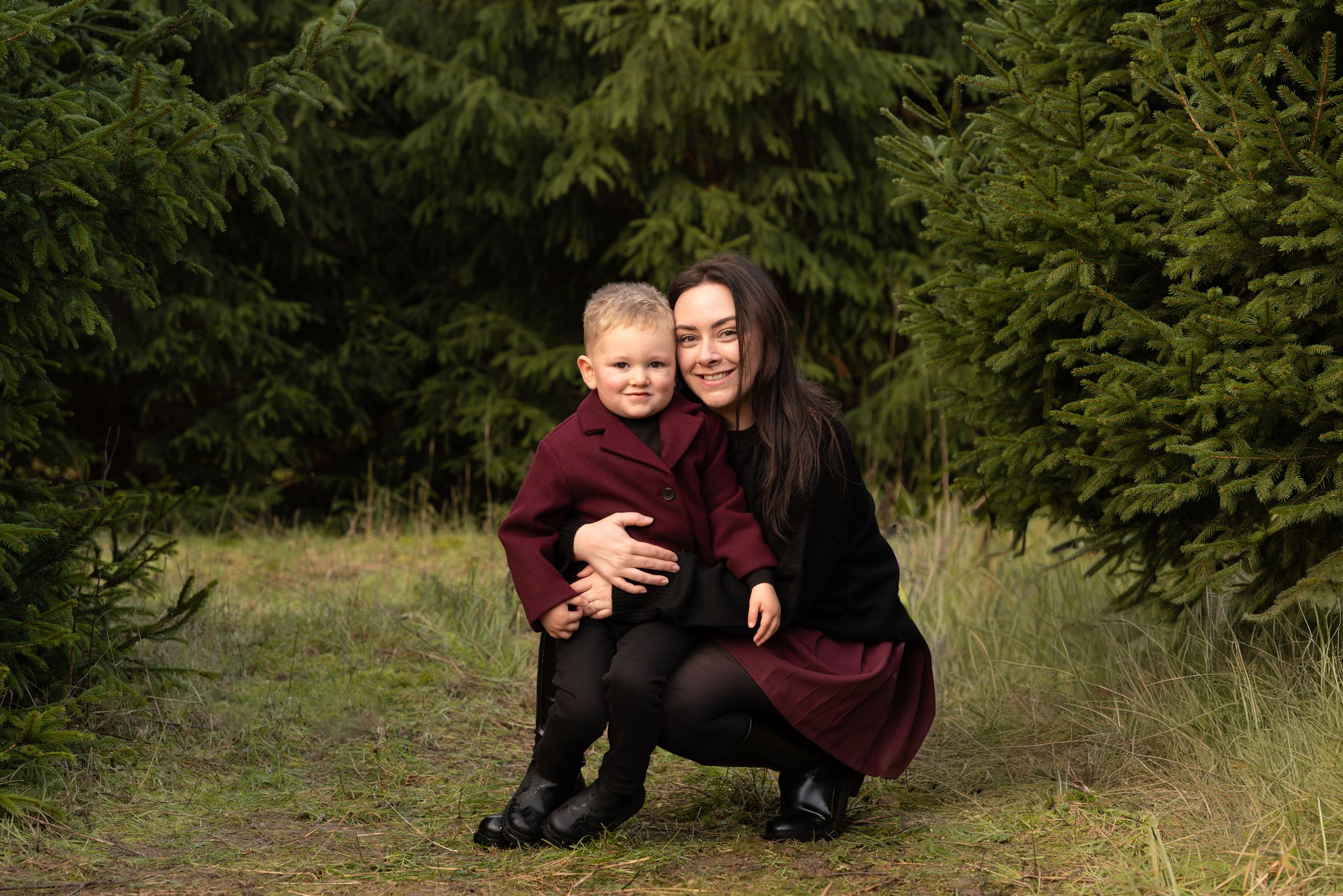 Smiling mother and young son in matching burgundy outfits crouch together on a grassy forest clearing surrounded by evergreen trees.