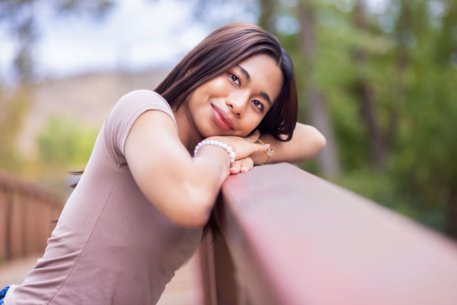 Young woman smiling and leaning on a wooden railing outdoors, with green trees in the background.