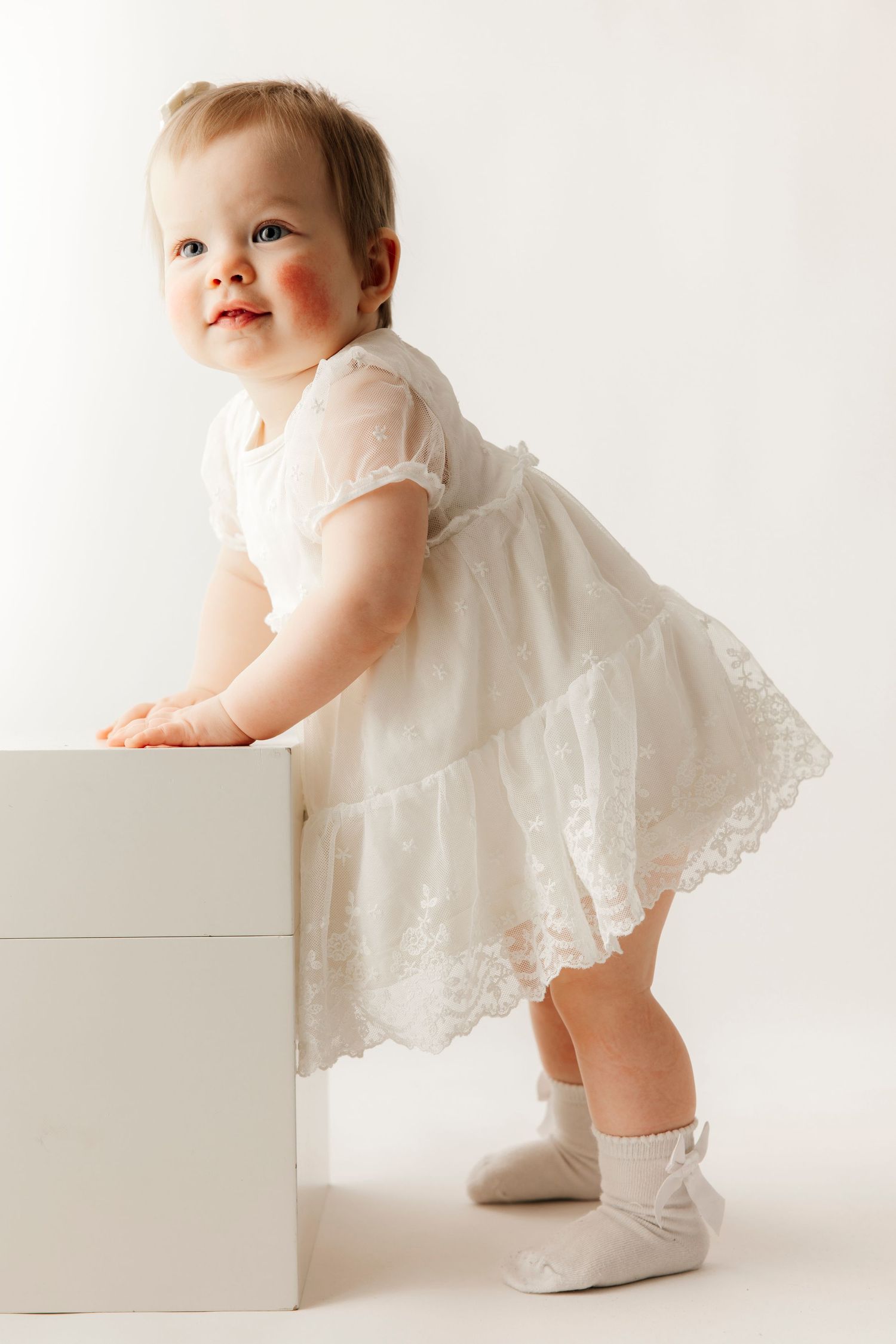 A cute baby in a white lace dress sits on the floor, touching her face near a small cake on a wooden stand.