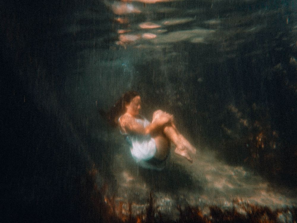 woman in a comforting pose under water in grainy film like abstract portrait taken during The Tide of Her underwater empowerment session NSW