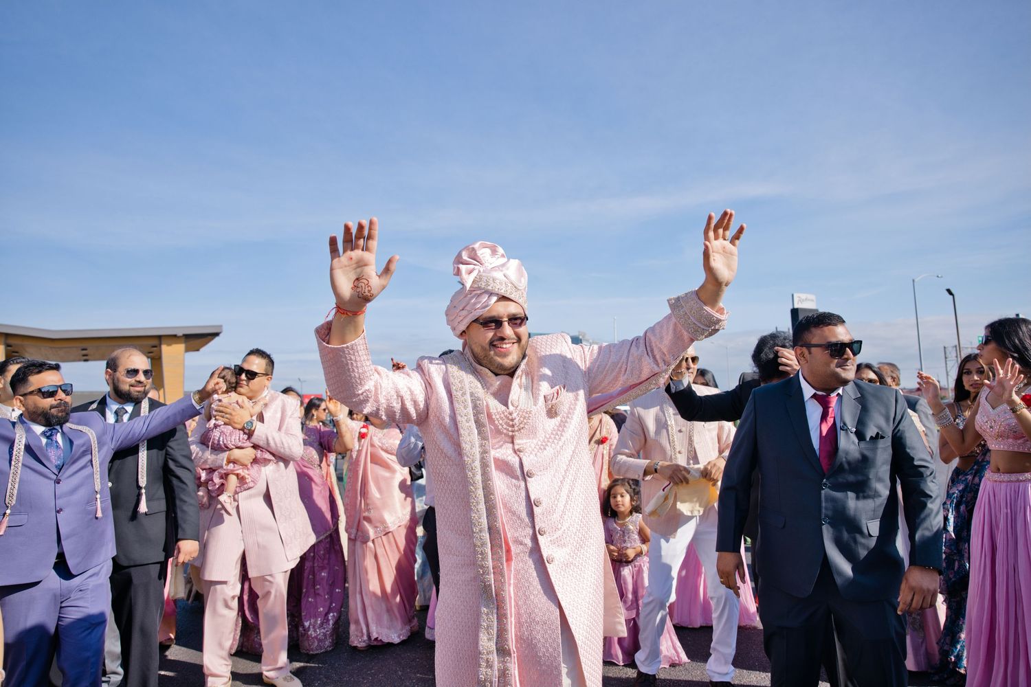 Groom in a pink sherwani and turban raises his hands and dances joyfully with wedding guests during a lively Indian baraat procession.