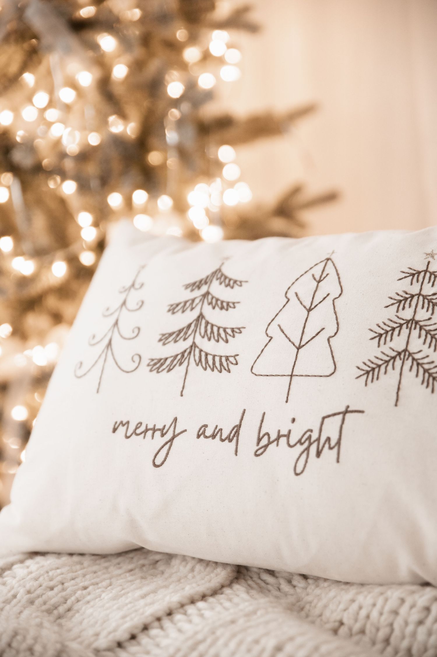 White decorative throw pillows with embroidered Christmas tree designs rest on a cozy knit blanket with twinkling lights in background.