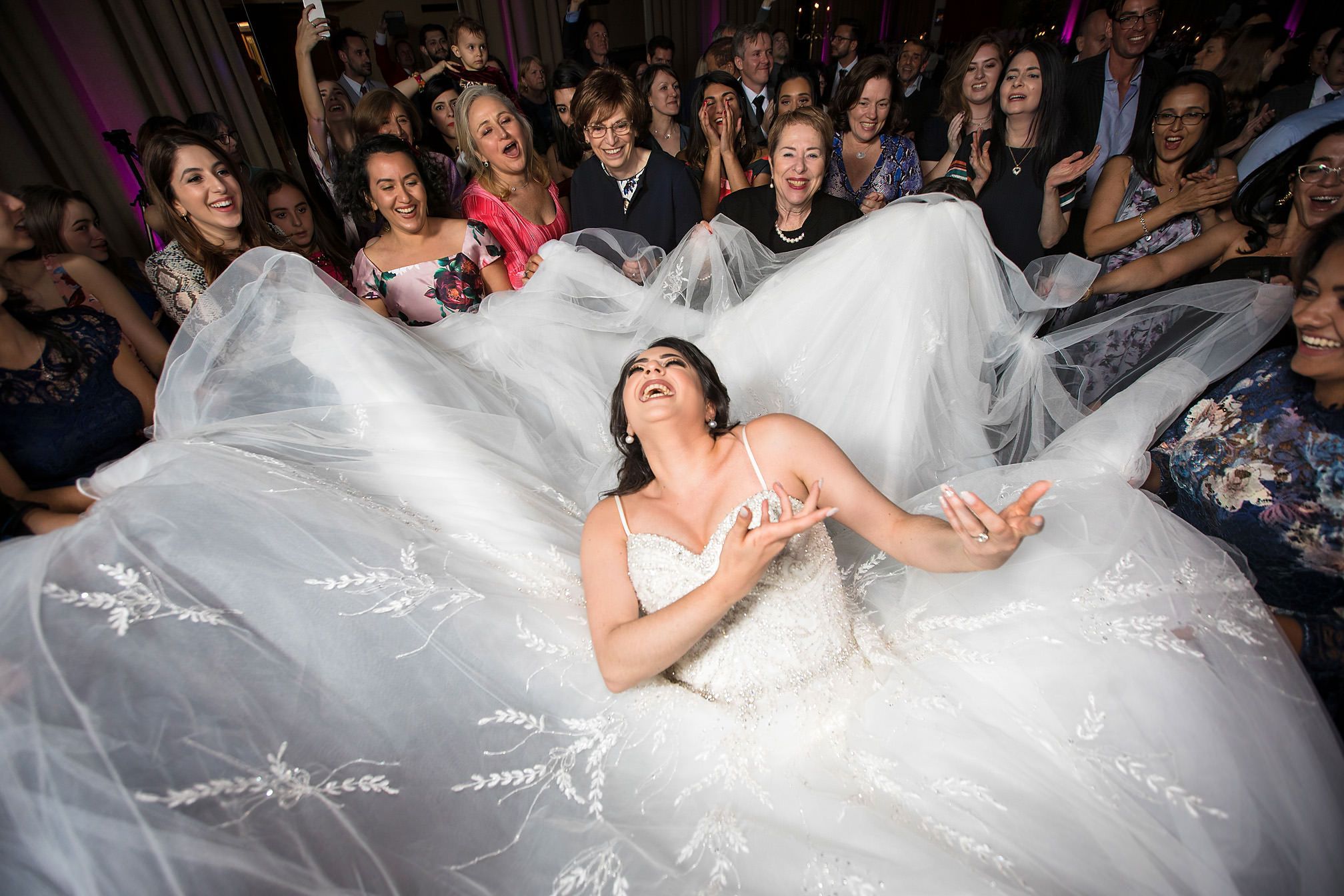 Bride in a voluminous white gown laughs joyfully on the dance floor surrounded by cheering wedding guests.